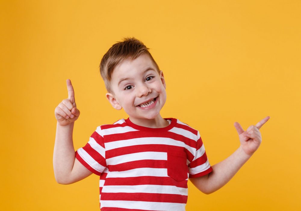 Smiling happy boy pointing fingers up at copyspace isolated over orange background